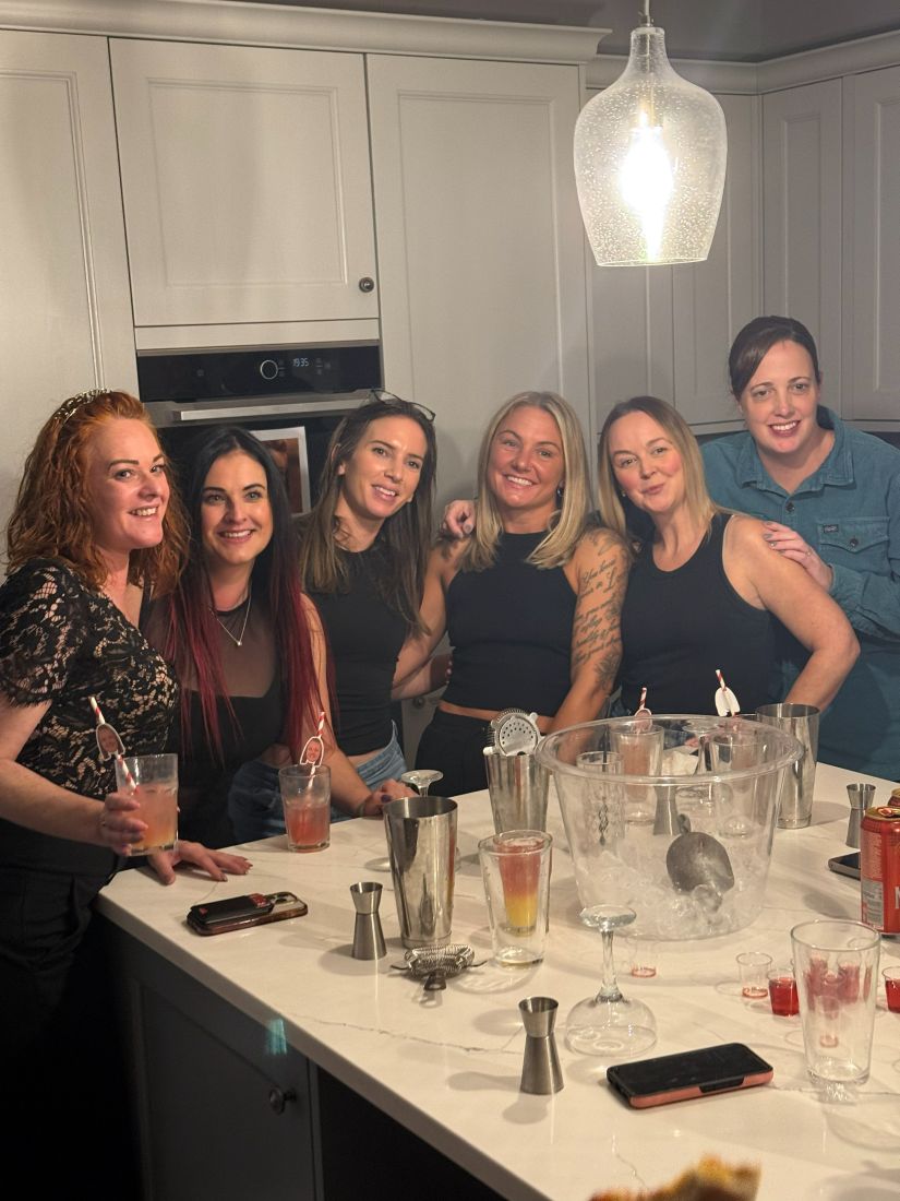 Six friends smiling with their finished cocktails around a kitchen island